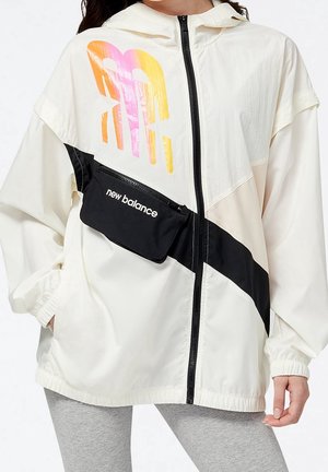 Kobieta nosząca białą kurtkę z kapturem na zamek błyskawiczny z czarnym ukośnym paskiem i dołączoną czarną saszetką New Balance, w połączeniu z szarymi legginsami.
