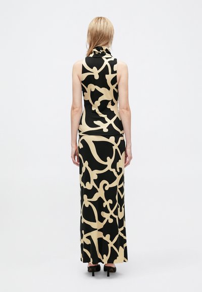 Mulher vestindo um vestido maxi sem mangas, com um padrão abstrato em preto e bege e um pescoço alto, de pé contra um fundo branco liso.
