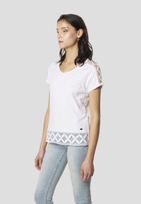 Biele bavlnené tričko so ženským výstrihom do V, krátkymi rukávmi a čipkovými detailmi pozdĺž lemu a na ramenách. Kombinované so svetlomodrými džínsami.