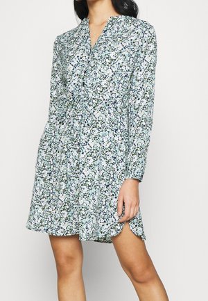 Femme portant une robe à manches longues, longueur genou, avec un motif floral bleu et blanc et une taille froncée.