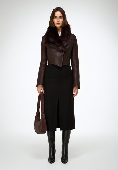 Giacca corta in pelle marrone con colletto in pelliccia, abbinata a una gonna midi nera con spacco frontale. La modella indossa stivali neri fino al ginocchio e una borsa marrone.