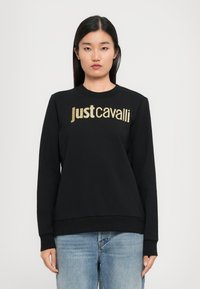 Donna che indossa un felpa nera con il logo dorato "Just Cavalli", abbinata a jeans azzurri, in piedi su uno sfondo chiaro e uniforme.