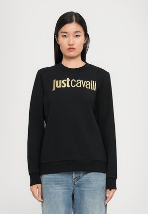 Frau trägt einen schwarzen Sweatshirt mit goldenen "Just Cavalli"-Logo, kombiniert mit hellblauen Jeans, stehend vor einem schlichten hellen Hintergrund.