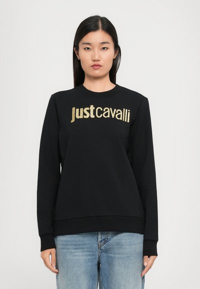 Femme portant un sweatshirt noir avec le logo doré "Just Cavalli", associé à un jean bleu clair, se tenant devant un fond clair.