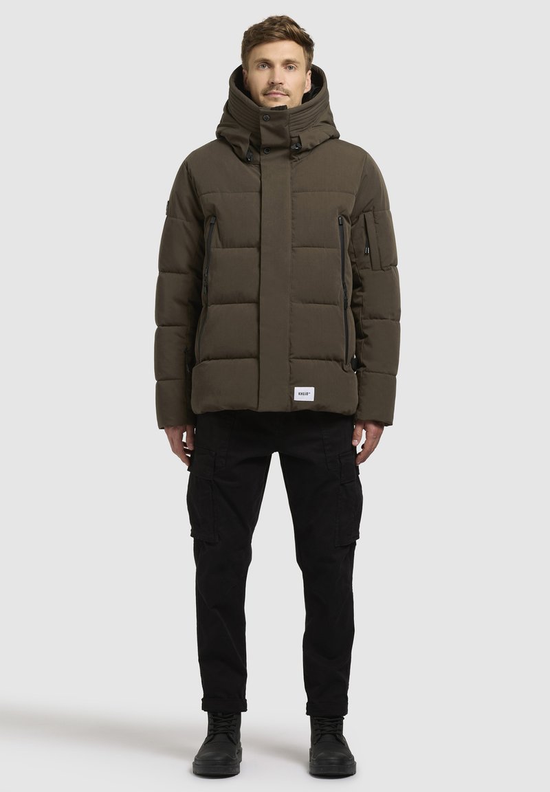 Padded olivgrüne Jacke mit hohem Kragen, mehreren Reißverschlusstaschen und verstellbare Kapuze. Getragen über schwarzen Cargo-Hosen und Stiefeln.
