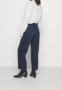 Osoba oblečená v tmavě modrých širokých džínách s vysokým pasem, světle šedém oversized svetru a černých kožených kotníkových botách, stojící na bílém pozadí.