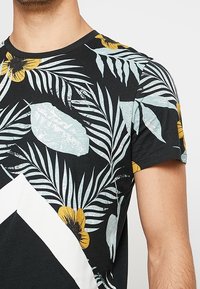 Černé tričko s žlutými květinami a světlomodrými tropickými listovými vzory, které nosí osoba ukazující horní část trupu a ruku.