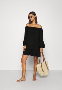 Vestito nero off-the-shoulder con maniche a campana e orlo arricciato, abbinato a una borsa di paglia intrecciata con manici in pelle marrone.