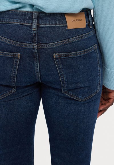 Dark blue denim jeans with a smooth texture, feature two back pockets, contrasting stitching, and a tan leather logo patch on the waistband.
