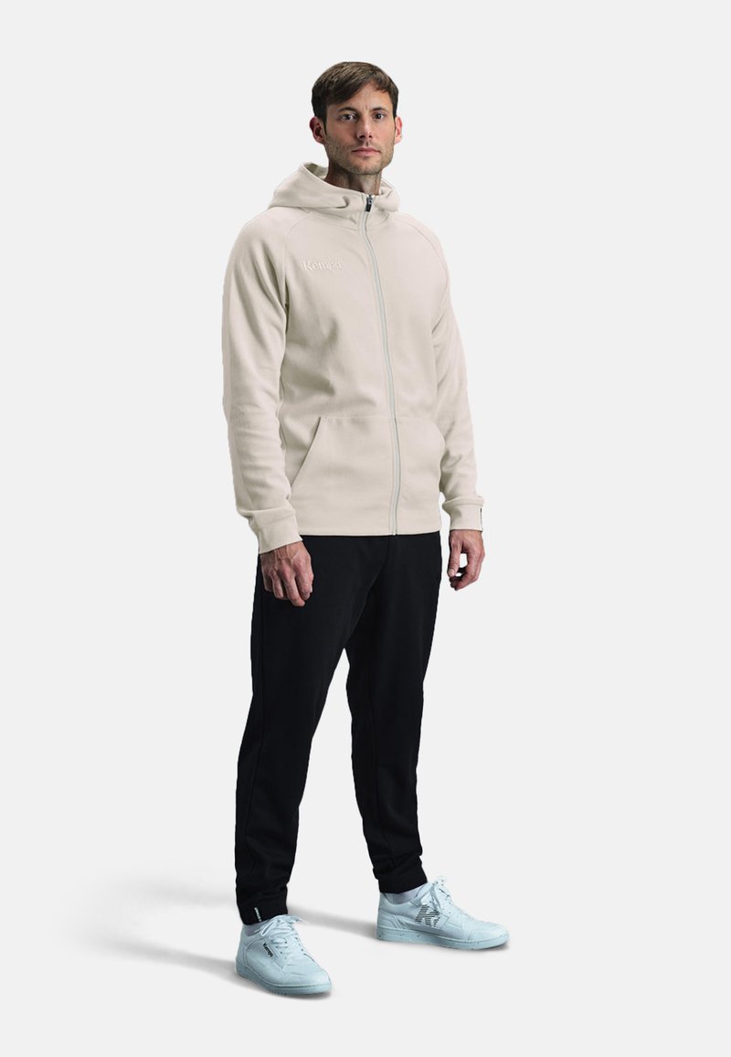 Lichtbeige rits-hoodie met hoge kraag, gecombineerd met zwarte broek. Heeft een gladde textuur en witte sneakers.