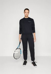 Conjunto preto de parte superior e calças em tecido de felpa com punhos canelados. Apresenta uma impressão do logótipo em verde. O modelo segura uma raquete de ténis azul e branca.