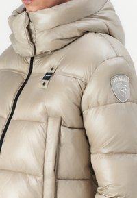 Beige pufferjack met een hoge kraag, zwarte rits en zijzakken. Het heeft een logopatch op de mouw en een glanzende, gestikte textuur.