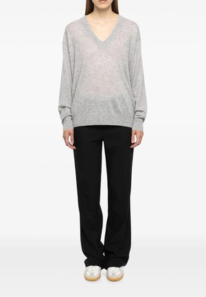 Donna con maglione grigio chiaro a V manica lunga, pantaloni neri larghi e scarpe da ginnastica bianche, in piedi davanti a uno sfondo neutro.