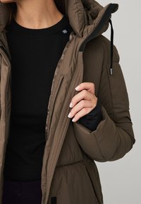 Brun puffjakke med lynlås og justerbar hætte, båret over en sort langærmet bluse, med kviltet tekstur og elastiske manchetter.