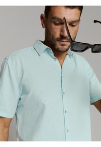 Man in een lichtblauw kortarm overhemd houdt bruine schildpadbril bij zijn gezicht tegen een neutrale achtergrond.