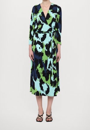 Frau trägt ein langärmliges Midi-Wickelkleid mit einem abstrakten Muster in Blau, Grün und Schwarz, kombiniert mit schwarzen Riemchen-High Heels.