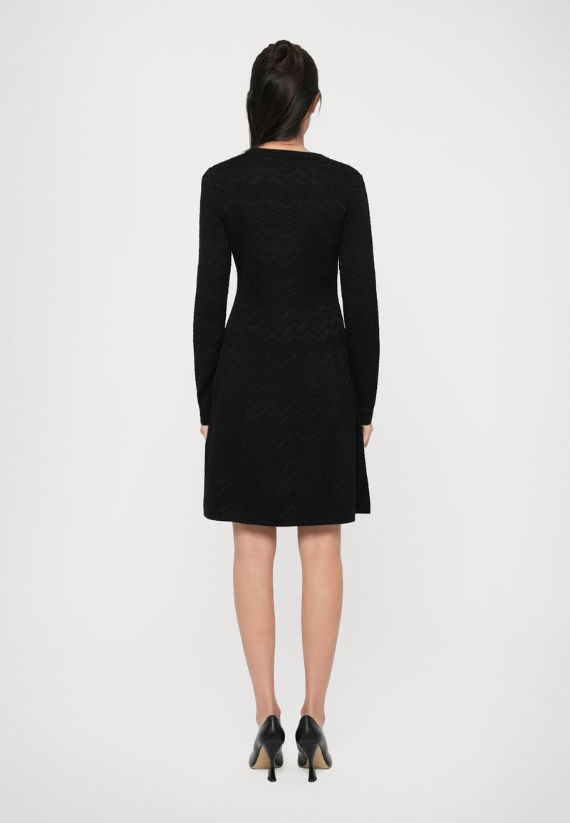 Robe noire à manches longues avec un motif en zigzag texturé, corsage ajusté, jupe évasée et col rond, associée à des chaussures à talons noires.