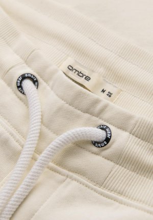 Jogging beige clair avec ceinture côtelée, cordon blanc et œillets noirs. Étiquette étiquetée "ombre" en taille M, détails de couture visibles.