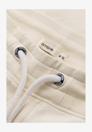 Jogging beige clair avec ceinture côtelée, cordon blanc et œillets noirs. Étiquette étiquetée "ombre" en taille M, détails de couture visibles.