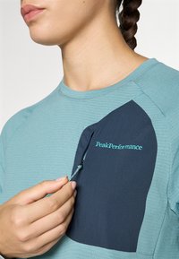 Himmelblaues, strukturiertes Langarmoberteil mit einer marineblauen Tasche mit Reißverschluss, akzentuiert durch ein türkisfarbenes "PeakPerformance"-Logo.