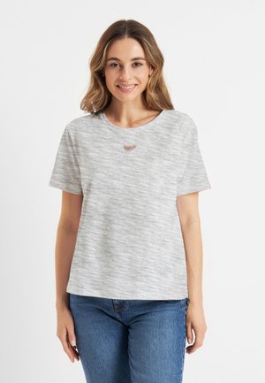 Jonge vrouw die glimlacht, gekleed in een lichtgrijze T-shirt met korte mouwen met een klein hartvormig uitsnijden bij de halslijn en blauwe jeans.