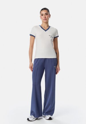 Naine seisab, seljas valge V-kaelusega T-särk siniste äärtega ja sinised laiade säärega püksid, valgete tossudega, valgel taustal.