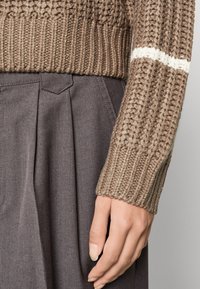 Brązowy sweter z dzianiny o ribbowanej fakturze, z białym paskiem. W połączeniu z ciemnoszarymi spodniami w plisy. Zbliżenie na rękaw i dłoń.