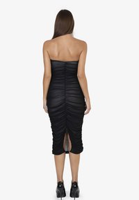 Abito nero senza spalline con tessuto arricciato, silhouette aderente, spacco sul retro e orlo fino al ginocchio, abbinato a tacchi neri lucidi.