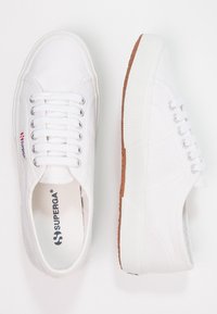 Para białych płóciennych trampków z białymi sznurówkami, srebrnymi oczkami, gumowymi podeszwami, widocznym logo marki "Superga" z boku i na wkładce.
