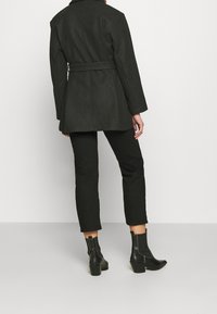 Cappotto in misto lana nera con vita abbottonata, colletto e maniche larghe, abbinato a pantaloni neri attillati e stivaletti con tacco neri a altezza caviglia.