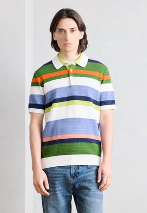 Νεαρός άνδρας με μεσαίου μήκους καστανά μαλλιά, φορώντας πολύχρωμο ριγέ πόλο και μπλε τζιν, στέκεται μπροστά σε λευκό απλό τοίχο.