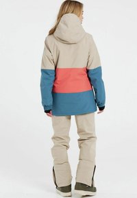 Skijacke in Beige, Blau und Koralle mit Kapuze, elastischen Bündchen und wasserdichtem Material. Bequeme Passform mit separaten Hosenbeinen und Reißverschlüssen.