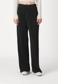 Pantalons noirs à plis, à jambe large avec une taille élastique, présentant un tissu texturé qui crée des lignes verticales. Portés avec des chaussures de couleur claire.