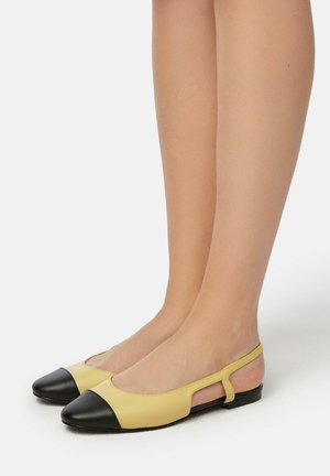 Fødder iført gule slingback-flats med sorte spidse tåkapper mod en ensfarvet hvid baggrund.