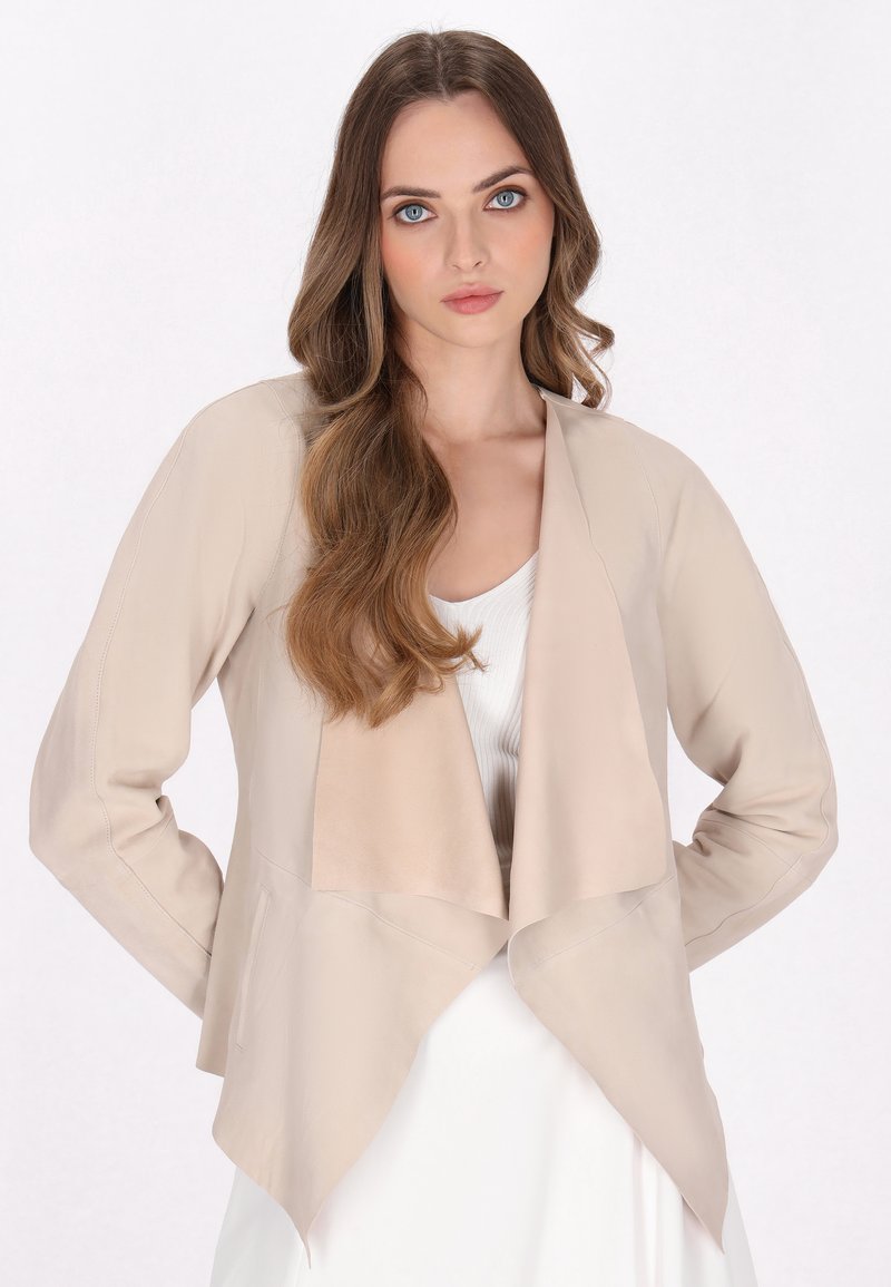 Beige faux læderjakke med åben front, draperet design, lange ærmer og en glat tekstur. Båret over en hvid top.