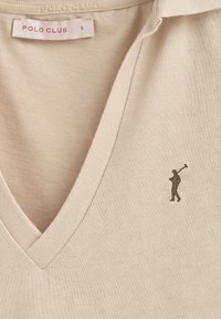 Beżowa koszulka polo z dekoltem w serek. Posiada małe logo przedstawiające postać trzymającą kij golfowy na lewej piersi. Gładki materiał bawełniany.