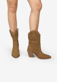 Botas de vaquero de cuero marrón claro con diseño perforado, punta afilada y tacón apilado. Presentan costuras en contraste y lengüetas de tirón en los laterales.