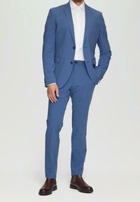 Niebieski garnitur o dopasowanym kroju, jednozgłowy żakiet, klapy w kształcie notcha i spodnie o prostym kroju. Noszony z białą koszulą i brązowymi botkami.