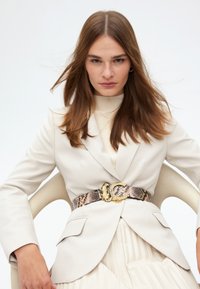 Beige kavaj över en krämfärgad tröja, ihop med ett texturerat bälte i ormtryck med en guldormsformad spänne. Plisserad kjol synlig nedanför.