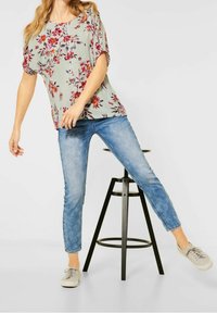 Blus med blommönster i ljusgrön tyg med lös passform, matchad med ljusblå jeans. Modellen bär ljusgrå sneakers och poserar nära en pall.