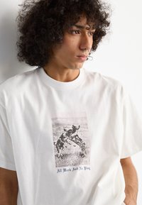 Hvid bomulds t-shirt med et centralt grafisk motiv af hunde, der leger, og teksten "Alt arbejde og ingen leg" i blå. Blød tekstur og afslappet pasform.