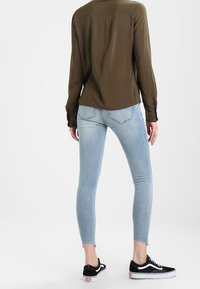 Camisa de manga larga verde oliva combinada con jeans ajustados azul claro y zapatillas negras. Los jeans tienen un dobladillo sin acabar y un diseño ajustado.