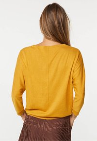 Keltainen, pitkähihainen neulepaita, jossa on leveä kaula-aukko ja suora takasauma, yhdistettynä ruskeisiin, teksturoituihin housuihin. Pehmeä materiaali, rento istuvuus.