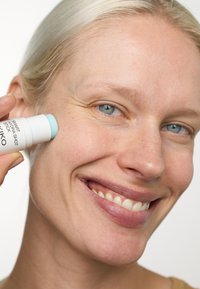 Stick Smart Hydra Shot, con involucro bianco e punta verde menta, applicato sulla guancia. Ingrandimento della texture della pelle e di un volto sorridente.