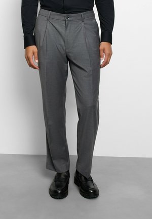 Man draagt grijze geplooide broek, zwart overhemd en zwarte leren schoenen, staand op een grijze vloer.