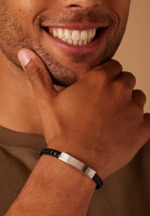 Zwarte gevlochten leren armband met een plat zilveren metalen plaatje. De pols is naakt behalve de armband, wat een gladde textuur toont.