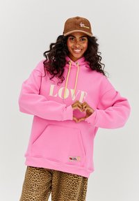 Růžová mikina s nápisem "LOVE" tištěným velkými krémovými písmeny, má přední kapsu a žluté tkaničky, v kombinaci s kalhotami s leopardím potiskem.