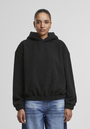 Sort fleece hoodie lavet af et tekstureret, fluffy materiale. Har en hætte med snoretræk og elastisk kant. Sammen med blå denim jeans.