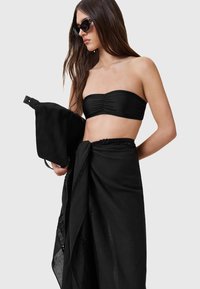 Top bandeau nero con dettagli arricciati, abbinato a un lungo sarong nero trasparente con intricate rifiniture in pizzo. Modella che tiene una borsa nera.