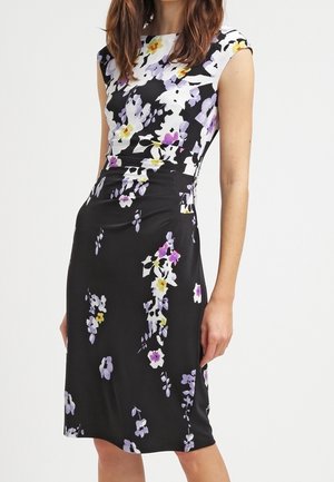 Robe noire sans manches avec imprimé floral blanc, jaune et violet, taille cintrée et ourlet longueur genou portée par une femme debout.